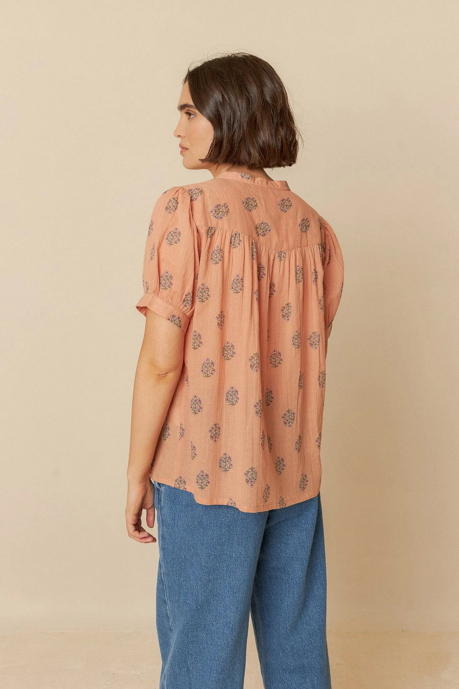 Woman wearing a patterned blouse and blue jeans against a beige background