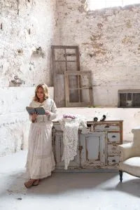 Woman standing in a rustic room with a distressed wooden cabinet and brick walls.